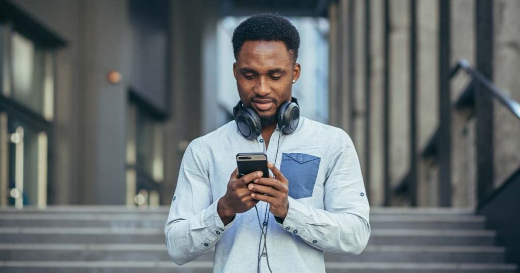 Young african american student man listens to podcast using app on phone, and big earphones