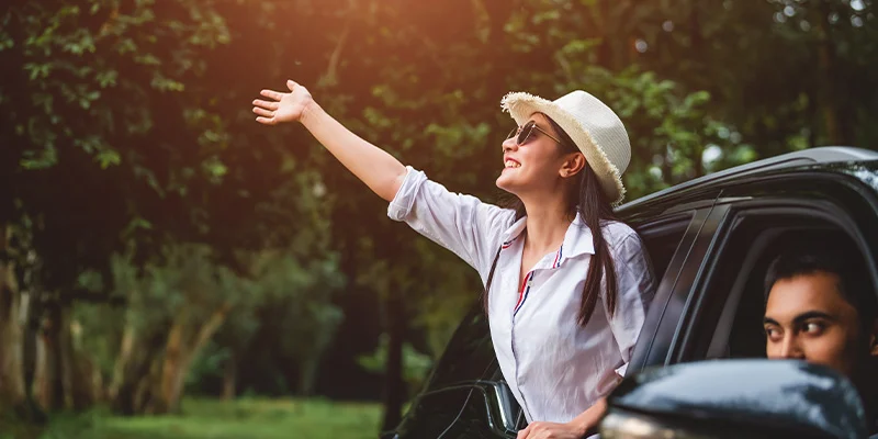 Happy woman waving hand outside open window car with her boyfriend on forest background