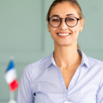 Smiling woman with glasses holding a book, in a professional setting with a French flag in the background
