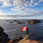 Adventurous woman holding a Canadian Flag on a Rocky Atlantic Ocean Coast during a sunny day. Taken in Sleepy Cove, Crow Head, Twillingate, Newfoundland, Canada.
