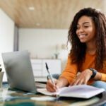 Note Taking Concept. Smiling young African American woman sitting at desk working on laptop and writing letter in paper notebook, holding pen and looking at screen. Happy millennial female using pc