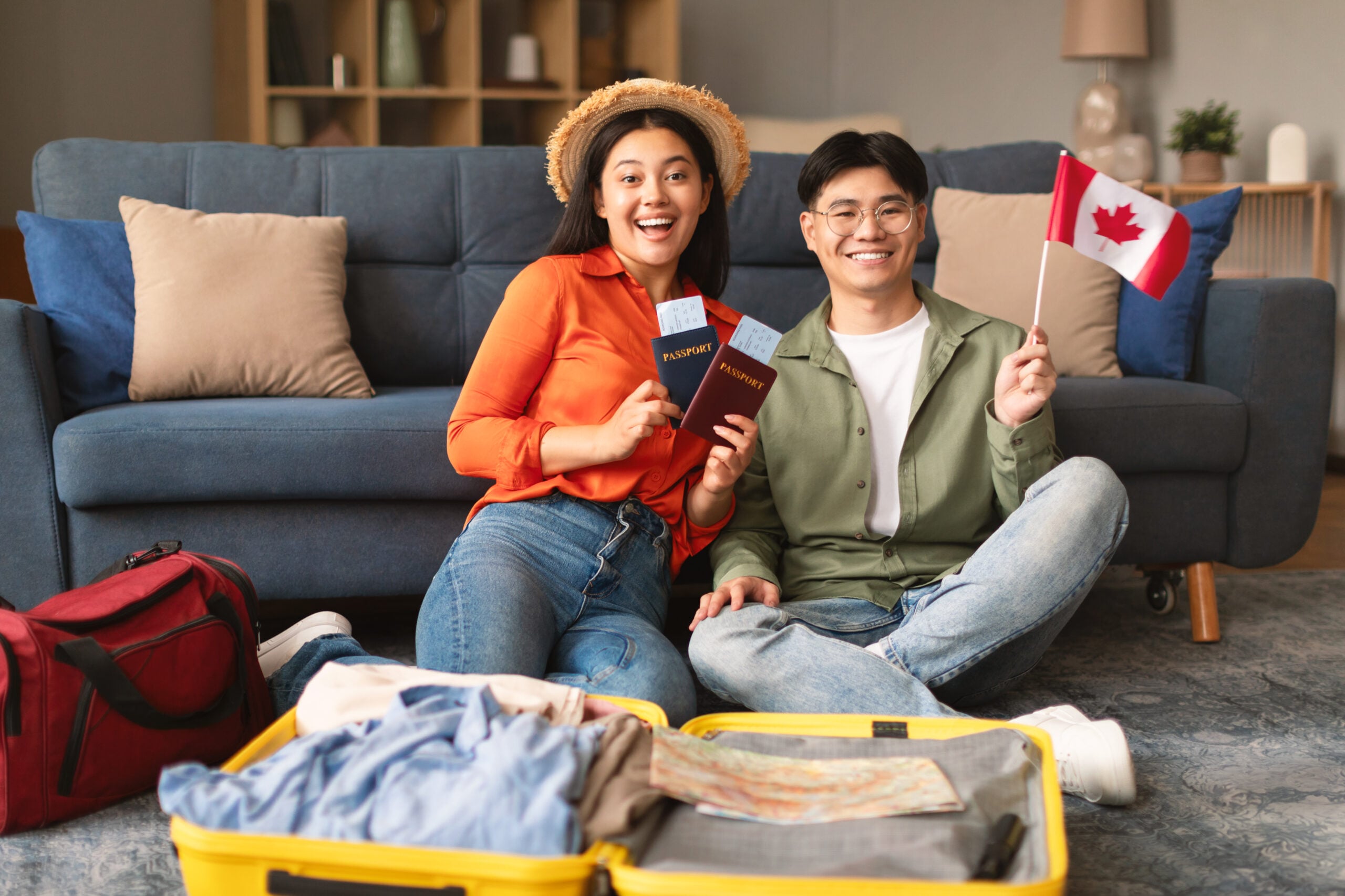 Excited Asian Young Couple Posing Holding Flag Of Canada, Packing Travel Suitcase For New Journey At Home. Spouses Smiling To Camera Preparing For Relocation And Trip Abroad