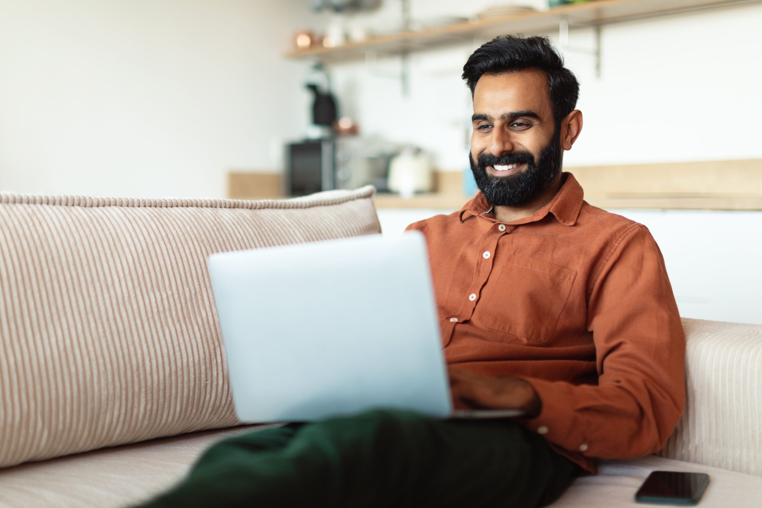 Freelance Concept. Indian Man Working Online At Laptop Computer Sitting On Couch At Home. Bearded Arab Business Guy Websurfing In Modern Living Room Indoors. Distance Job And Internet Technology