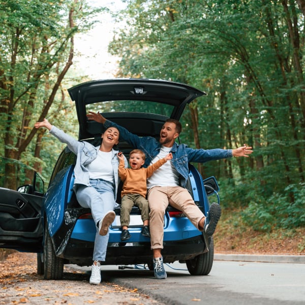 Happy couple with a young child sitting in the trunk of a vehicle in a leafy laneway, celebrating with their arms in the air.