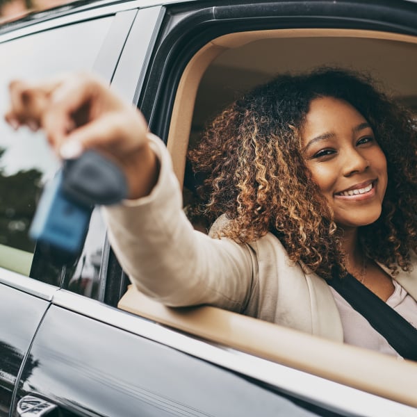 Young woman with dark curly hair sitting in the front seat of a car with the window open and her hand holding her keys in the photograph foreground