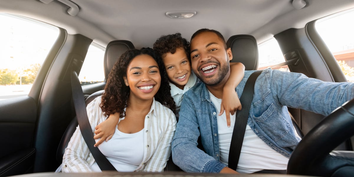 Young happy couple sitting in the front seat of a vehicle with seatbelts on with a child in the back seat hugging them.