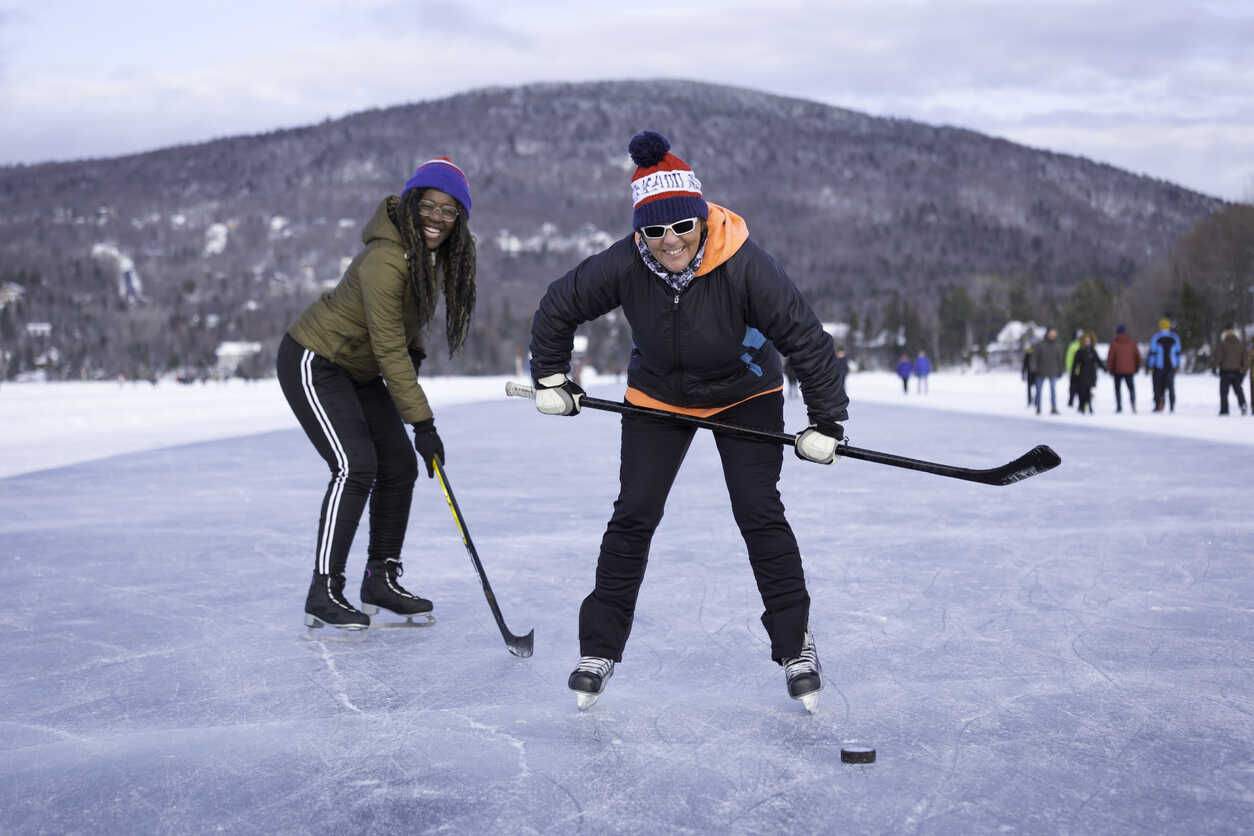 Two women skating on ice in Canada with hockey sticks and a puck, and a group of people looking on