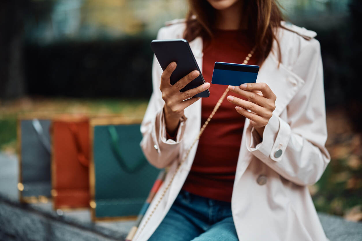 Photograph of a woman wearing a seasonal coat and sitting outdoors holding up her phone and credit card for credit monitoring. She has a series of shopping bags in the background of the photo