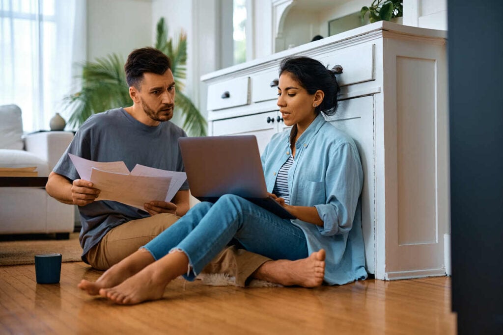 Couple sitting on the floor looking at budgeting spreadsheets on a laptop and some paper bills