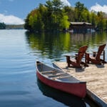 Two Adirondack chairs on a wooden dock overlook the serene blue waters of a Muskoka lake in Ontario, Canada. Nearby, a red canoe is tied to the pier, alongside life jackets and a paddle. Space for text.