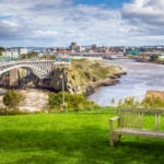Photo of Saint John, New Brunswick, from a park near the Reversing Falls Bridge. An empty wooden bench is in Foreground.